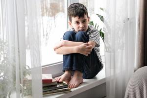 A sad boy sits on the windowsill hugging his knees. Bad mood, depression. The boy is sad alone at home photo