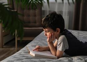 un de seis años chico es leyendo un libro con entusiasmo, acostado en el cama. hogar enseñanza. preparando un niño para colegio foto