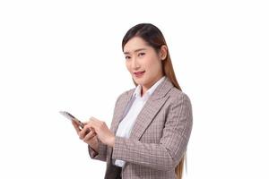 profesional asiático trabajando mujer en un blanco camisa y a rayas traje en pie con confianza sonriente y mirando a el cámara es haciendo inteligente teléfono a cheque información aislado en blanco antecedentes. foto