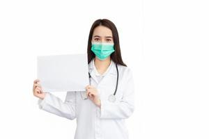 Asian woman professional doctor who wears medical coat and face mask stands and shows white paper to say something isolated on white background in Coronavirus healthcare concept. photo