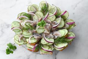 salado arenque con especias, Fresco pepino, hierbas y cebollas en un plato en un ligero antecedentes. en escabeche rebanado pescado con un pan. foto