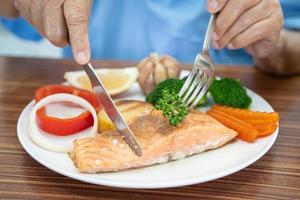 Asian senior or elderly old lady woman patient eating Salmon steak breakfast with vegetable healthy food while sitting and hungry on bed in hospital. photo