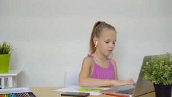 Serious schoolgirl sitting at table with laptop and textbook and doing homework. video