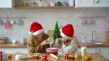 Little girls making Christmas gingerbread house at fireplace in decorated living room.