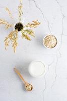 Oat milk in a glass, oatmeal in a bowl and ears in a jug on a light table. Alternative plant food. Vertical view. photo