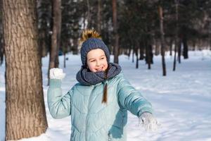 A cheerful girl in warm clothes throws a snowball in a winter park. Outdoor games. photo