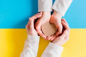 The concept is no war in Ukraine. Children's and women's hands hold a cardboard heart in their palms against the background of the Ukrainian flag. Top view. photo