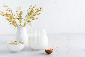 Oat milk in a glass, oatmeal in a bowl and ears in a jug on a light table. Alternative plant food photo