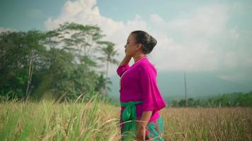 uma mulher asiática apreciando a vista enquanto colhe o campo de arroz em um vestido rosa e lenço verde em seu corpo na aldeia video