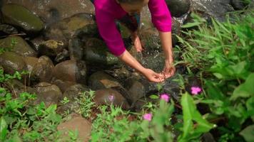 uma lindo ásia mulher lavagens dela mãos dentro a bambu chuveiro perto a rio enquanto vestindo uma Rosa vestir com verde folhas e arbustos video