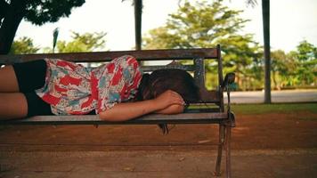 an Asian woman lying on a park bench under a large shady tree alone video