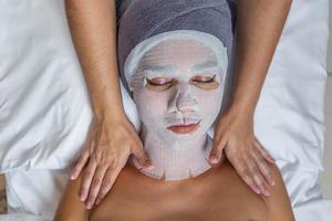 Woman with her face covered with gauze for skin treatment receiving shoulders massage by beautician photo