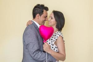 Cheerful Hispanic couple with heart shaped balloon hugging in studio photo