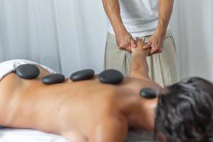 frente lado ver de un masajista haciendo un mano y brazo masaje a adulto mujer con caliente piedras en su espalda para masaje espalda tratamiento en spa salón foto