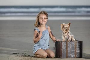 alegre niña cerca maleta y perro en arenoso playa foto