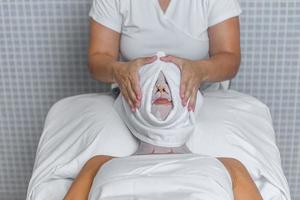 Beautician applying a towel to the face of a woman who is covered with gauze for skin treatment photo