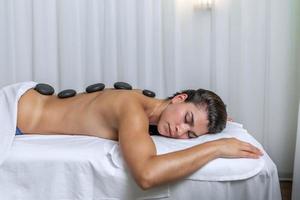 Side view of a young woman lying face down in a massage table with spa stones on her back photo