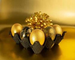 Five eggs, painted in gold and black, lie in a dark textured stand on a shiny background. The holiday of light Easter photo