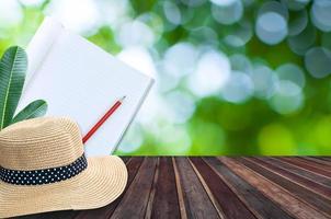 Notebook with pencil and straw hat on wooden table with green bokeh background photo