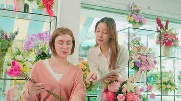 Two young beautiful female florist partners discussing bunch of blossoms arrangement, decorating with lovely ribbons, happy work in colorful flower shop store with blooms, SME business entrepreneur. video