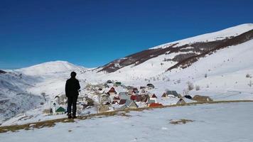 Aerial drone view of a solo hiker admiring a mountain village during winter. Snow white landscape and mountaineer lifestyle. video
