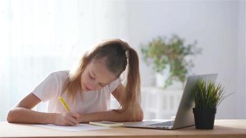 Serious schoolgirl sitting at table with laptop and textbook and doing homework. video