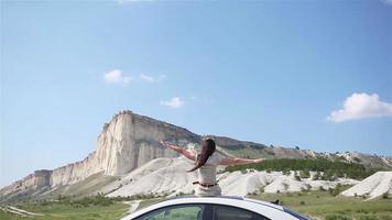 Happy girl sitting on roof of her car, relaxing and enjoying road trip video