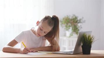 Serious schoolgirl sitting at table with laptop and textbook and doing homework. video