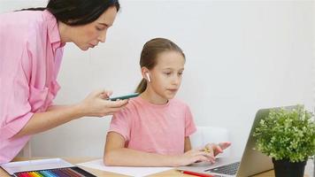 schoolmeisje zittend Bij tafel met laptop en leerboek en aan het doen huiswerk terwijl jong moeder helpen haar video