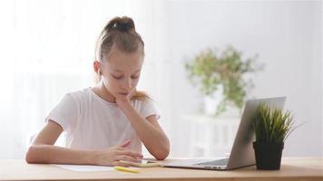 Serious schoolgirl sitting at table with laptop and textbook and doing homework. video