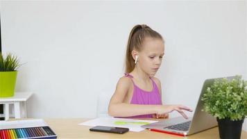 Serious schoolgirl sitting at table with laptop and textbook and doing homework. video