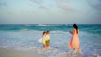 pequeño adorable muchachas y joven madre a tropical playa en calentar noche. mamá tomar un foto de hermosa niños en el puesta de sol video
