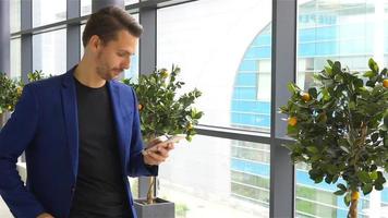 städtischer geschäftsmann, der innen am flughafen mit dem smartphone spricht. lässiger kleiner Junge mit Anzugjacke. kaukasischer mann mit handy am flughafen, während er auf das einsteigen wartet video