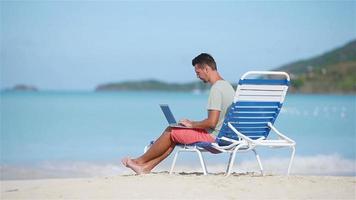 Young man with laptop on tropical caribbean beach. Man sitting on the sunbed with computer and working on the beach video