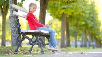 poco adorabile ragazza con smartphone nel autunno. ragazzo avendo divertimento a caldo soleggiato autunno giorno all'aperto video