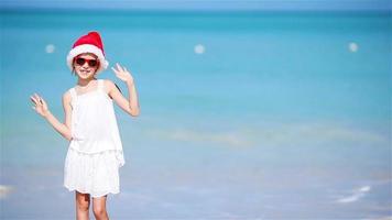 Little adorable girl in Christmas hat on white beach during Xmas vacation. Happy smiling kid looking at camera video