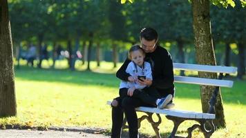 contento familia en caer. padre con teléfono inteligente y pequeño niño en otoño al aire libre video