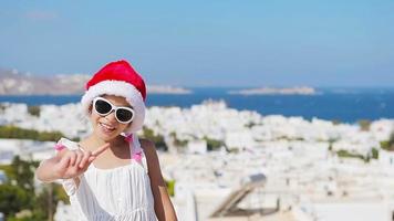 liten flicka i santa hatt utomhus bakgrund gammal gata på mykonos. unge på gata av typisk grekisk traditionell by med vit väggar och färgrik dörrar på mykonos ö, i grekland video