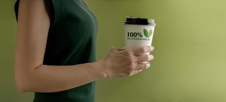 Biodegradable and Compostable Packaging Concept. Closeup of Woman Holding a Hot Cup of Coffee against a Green Wall. Zero Waste Materials. Environment Care, Reuse, Renewable for Sustainable Lifestyle photo