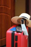 New normal concept. Close up view of a smartphone with an immune digital health passport, a red suitcase, protective mask, straw hat, passport and sunglasses. Traveling by plane during a pandemic. photo