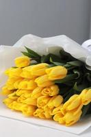 beautiful big bouquet of yellow tulips in a white package on white table and gray wall background close up. Congratulation. Festive background. copy space. photo