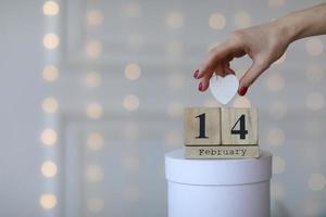 concepto de día de san valentín. fecha 14 de febrero en el calendario de cubos de madera en una caja de regalo blanca y corazón blanco en la mano. bokeh fondo dorado y blanco foto