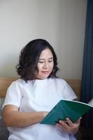 joven asiático embarazada mujer leyendo libro en el cama a hogar mientras participación su barriga foto