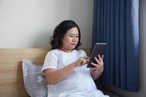 Young Asian Pregnant Woman using digital pen for drawing and taking note on her digital tablet at home near the window photo