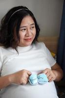 Photo of Happy Asian pregnant woman sitting in bed and touching her belly at home  With Small shoes for the unborn baby in the belly