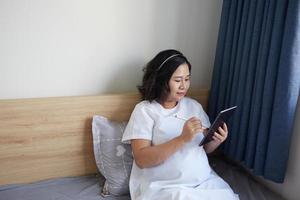 Young Asian Pregnant Woman using digital pen for drawing and taking note on her digital tablet at home near the window photo