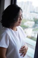 Photo of Happy Asian pregnant woman sitting in bed and touching her belly at home . With Small shoes for the unborn baby in the belly