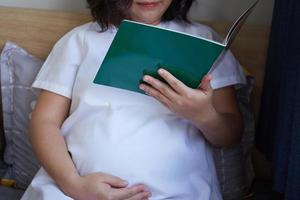 Young Asian Pregnant Woman reading book on the bed at home while holding her belly photo