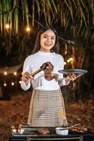 Portrait of Happy Asian young woman cooking meat on barbecue grill at new year party. Bar-B-Q or BBQ on traditional stove. Night Party, people and celebration concept. photo