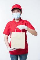Image of a happy young delivery man in red cap blank t-shirt uniform face mask gloves standing with empty brown craft paper packet isolated on light gray background studio photo